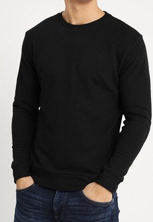 Mężczyzna ubrany w prostą czarną bluzę z długim rękawem, z rękami w kieszeniach, w połączeniu z niebieskimi dżinsami, na białym tle.