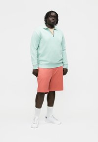 Moški stoji oblečen v mintasto zelen pulover s četrtinskim zadrgo, koralne kratke hlače, bele nogavice in bele superge na enobarvni belem ozadju.