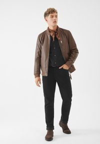 Giubbotto bomber in pelle marrone con polsini a coste, camicia nera button-down, pantaloni neri, sciarpa marrone e stivaletti marroni in un contesto neutro.