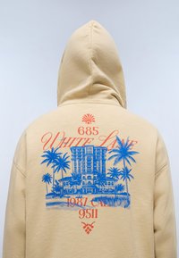 Sweat à capuche beige avec la capuche relevée, présentant un motif graphique bleu de palmiers et de bâtiments, ainsi qu'un texte rouge indiquant "685 White Lane 1987 Cali 9511" au dos.