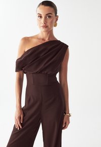 Rjava enodelna obleka z enim ramenom, z nagubanim materialom, pas prilagojen, široke hlače in gladka tekstura. Na zapestju videti zlati dodatek.