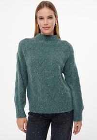 Groene gebreide trui met een hoge kraag, voorzien van een zigzagpatroon en zilveren spikkels, gecombineerd met donkere jeans versierd met kleine witte stippen.