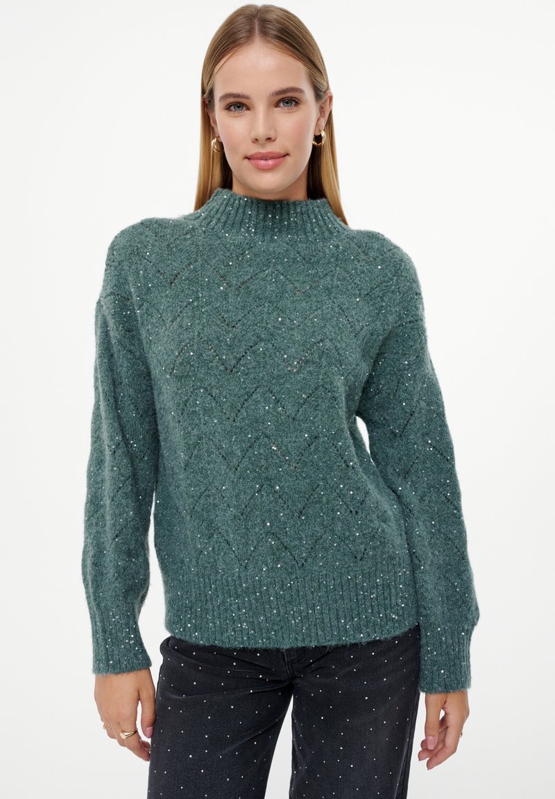 Groene gebreide trui met een hoge kraag, voorzien van een zigzagpatroon en zilveren spikkels, gecombineerd met donkere jeans versierd met kleine witte stippen.