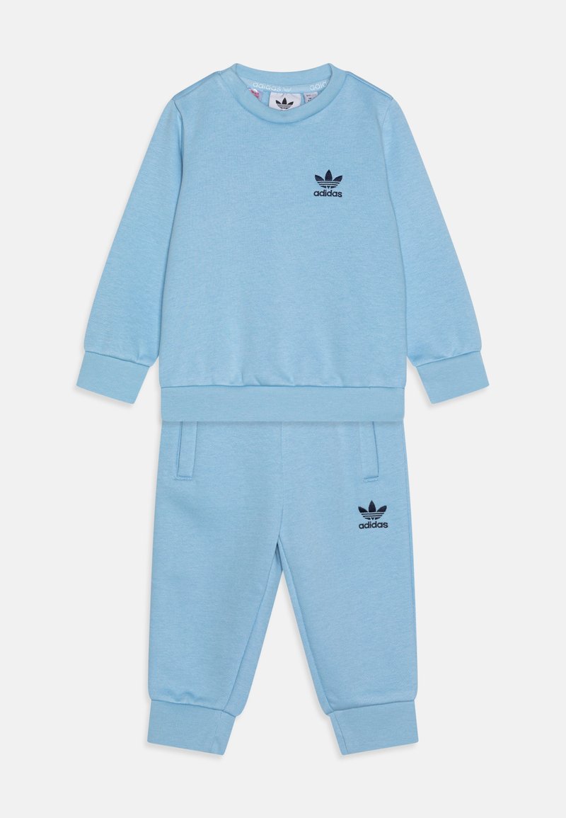 Šviesiai mėlynas medvilninis treningų kostiumas, turintis gikių rankoves, apvalią iškirptę ir juodą Adidas logotipą ant krūtinės bei šlaunies.