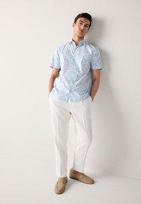 Rövid ujjú, világoskék virágmintás ing fehér és kék mintákkal, fehér nadrággal és bézs bebújós cipővel. Egyszínű háttér.