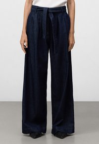 Vida byxor i mörk denim med bälte i midjan. Slät textur, rena linjer och sidofickor. Sömmen ligger ovanför skorna.