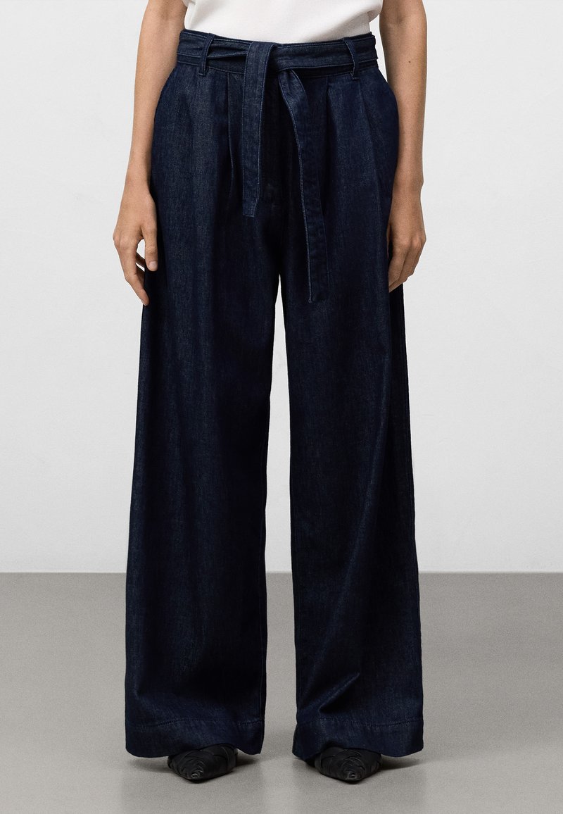 Vida byxor i mörk denim med bälte i midjan. Slät textur, rena linjer och sidofickor. Sömmen ligger ovanför skorna.