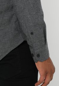 Grijs, gestructureerd T-shirt met lange mouwen en knoopsluitingen aan de manchetten. Zwarte knopen en contrasterende naden op de mouw. Zichtbaar gecombineerd met een zwarte broek.