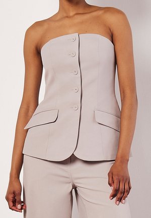 Haut beige sans bretelles, ajusté, avec boutons sur le devant et deux poches à rabat, associé à un pantalon beige assorti porté par une personne debout.