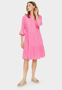Rochie roz, până la genunchi, cu mâneci până la trei sferturi și design în straturi. Fabricată dintr-un material moale, cu o croială lejeră. Purtată cu sandale crem cu talpă joasă.