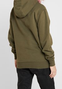 Sudadera verde oliva con capucha ajustable, puños y dobladillo acanalados. Presenta un corte relajado, hecha de un tejido suave, mostrada desde la parte de atrás.