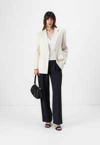 Beige skreddertilpasset blazer med to knapper, hvit v-hals topp, marineblå vide bukser, svart håndveske og svarte hæler.