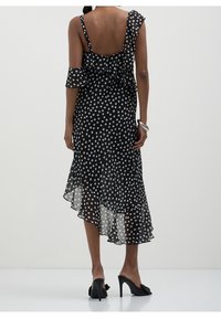 Robe noire à pois blancs en tissu transparent, avec des épaules volantées, un ourlet asymétrique et un dos nu. Portée avec des chaussures à talons hauts.