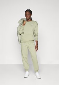Felpa verde chiaro con tasca frontale e logo, abbinata a pantaloni della tuta coordinati e sneakers bianche. Il modello tiene in mano un giubbotto imbottito.