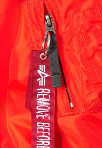 Röd nylonjacka med en dragkedjeficka, med en svart dragkedjeöppnare. En röd tygetikett med texten "REMOVE BEFORE" är fäst.