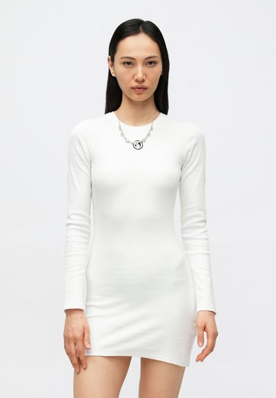 Witte jurk met lange mouwen, een aansluitend ontwerp en een geribbelde textuur. Zilveren ketting met een cirkelvormige hanger en kralenaccenten rond de hals gedragen.