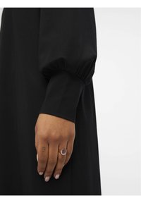 Schwarzes Kleid mit langen, puffigen Ärmeln. Nahaufnahme einer Hand, die einen Ring mit einem lila Stein trägt und ein Tattoo am Handgelenk zeigt.