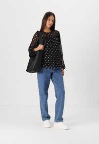 Blusa nera trasparente a maniche lunghe con pois, abbinata a jeans larghi blu chiaro e sneakers bianche. Una borsa nera è indossata a spalla.