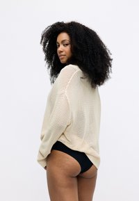 Beige, löst gestrickter Pullover in lockerer Passform mit weiten Ärmeln und einem strukturierten Muster. Kombiniert mit schwarzen Bikinihosen.