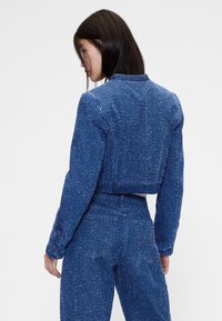 Modra jeans jakna do pasu z teksturirano končno obdelavo, dolgimi rokavi in zapenjanimi manšetami. Pogled od zadaj prikazuje oprijeto zasnovo z gladkim ovratnikom.