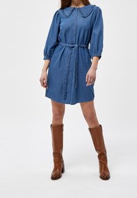 Jeansklänning med en volangkrage, puffärmar och bälte i midjan. Klänningen är knälång och bärs med höga bruna läderstövlar.