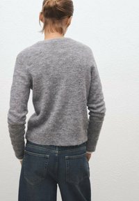 Siv svetar iz mehkega pletenine z okruglim izrezom in dolgimi rokavi, v kombinaciji s temno modrimi kavbojkami z zadnjimi žepi.