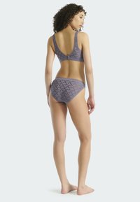 Žena stojící bosá, otočená zády, oblečená v odpovídajícím fialovém květinovém podprsenkovém a kalhotkovém setu na světlém jednolitém pozadí.