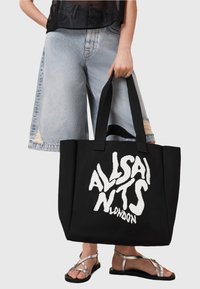 Une personne portant un short en denim clair et des sandales argentées tient un grand sac fourre-tout noir avec un texte blanc déformé "ALLSAINTS LONDON".