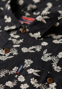Camisa corta de manga con estampado floral negro que presenta patrones botánicos blancos, botones marrones y una etiqueta de marca con acentos rojos y azules.