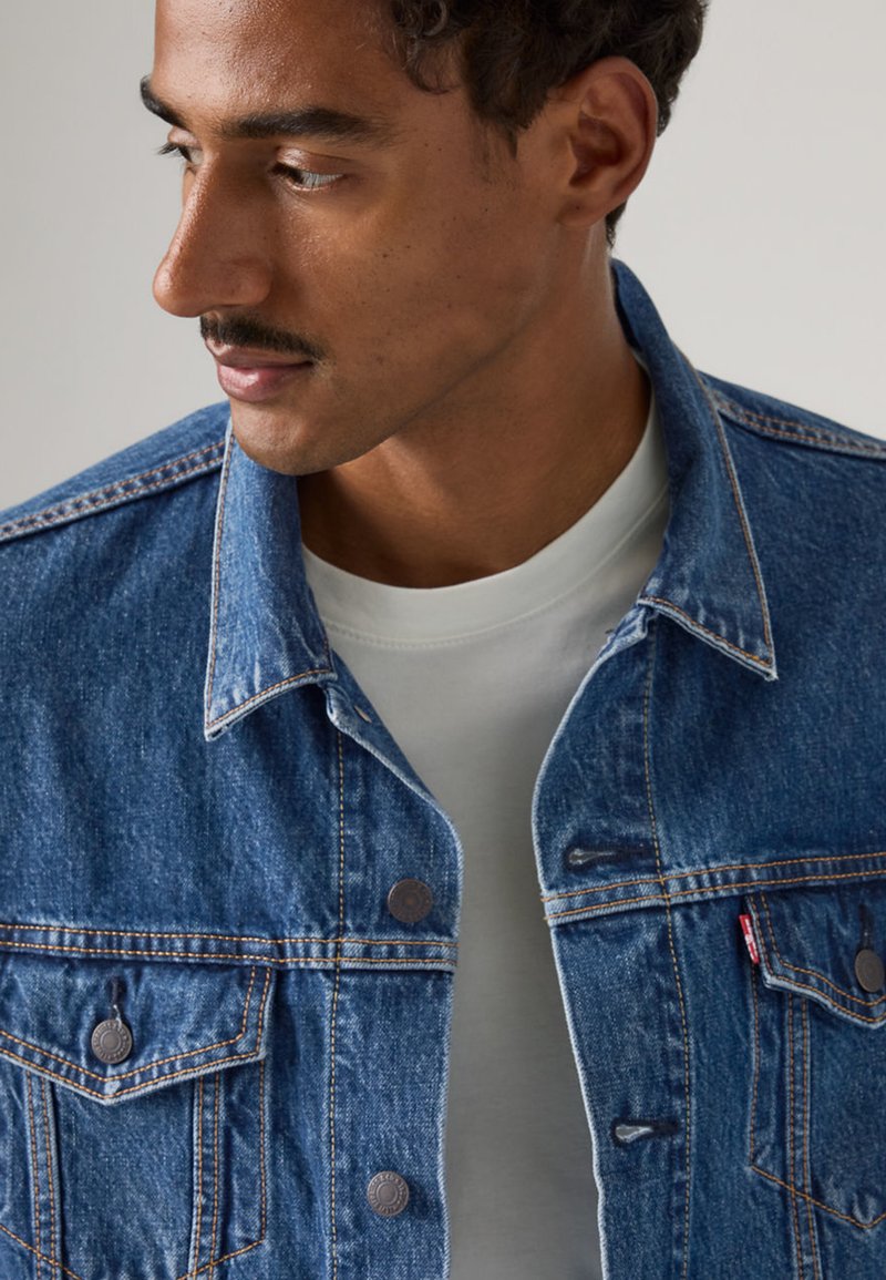 Blaue Jeansjacke mit Knopfverschluss, zwei Brusttaschen, kontrastierender Naht und klassischem Kragen, getragen über einem hellgrauen Hemd.