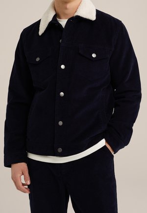 Homme portant une veste en velours côtelé bleu marine foncé avec col sherpa blanc et boutons-pression argentés, associée à un pantalon sombre assorti.