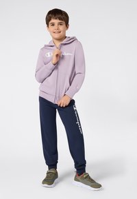 Chlapec oblečený v fialovom zipsovom mikine, námorníckych teplákach s bielym nápisom a olivovozelených teniskách, stojí na neutrálnom pozadí.