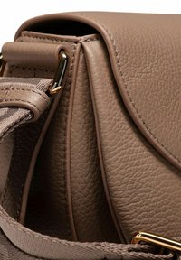 Primo piano di una borsa in pelle color tortora con trama, bordi cuciti, dettagli in oro e tracolla regolabile in tessuto passata attraverso un anello metallico.