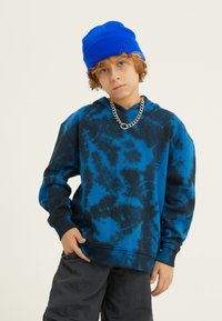 Felpa con cappuccio blu e nera in tie-dye, con tasca a marsupio, polsini a costine e cappuccio, abbinata a un berretto di lana blu e una collana in argento.