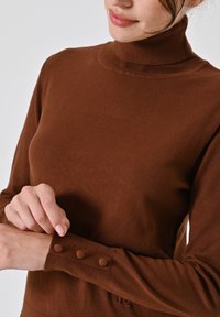 Brauner Rollkragenpullover aus weichem Material, mit Knopfdetails an den Manschetten und enganliegendem Design. Glatte Textur, schlicht und elegant.
