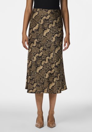 Frau trägt eine braun-schwarze Midirock mit Schlangenprint, ein schwarzes Oberteil und beigefarbene spitze High Heels, steht vor weißem Hintergrund.