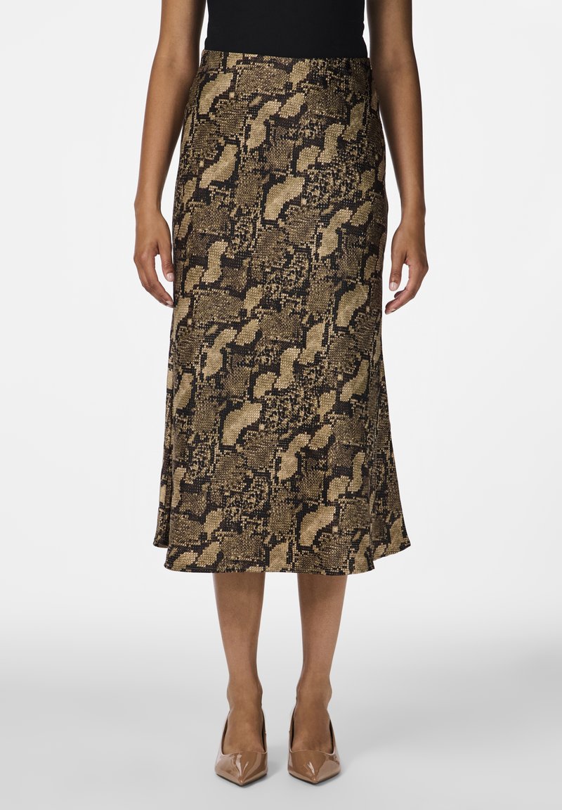 Femme portant une jupe midi imprimé serpent marron et noir avec un haut noir et des escarpins beiges à bout pointu, debout devant un fond blanc.