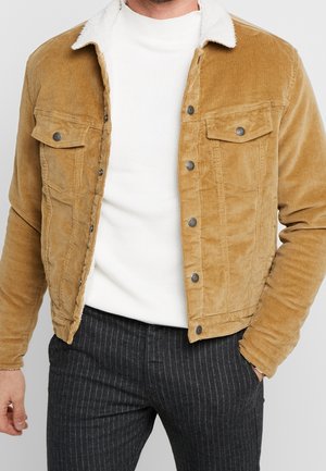 Mężczyzna ubrany w piaskową kurtkę ze sztruksu z białym kołnierzem z polaru, biały prążkowany sweter i ciemne spodnie w prążki, z ręką w kieszeni.