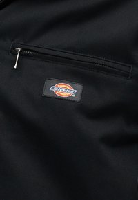 Tissu noir avec une poche zippée horizontale au-dessus d'un patch logo rectangulaire Dickies aux couleurs rouge, orange et bleu.