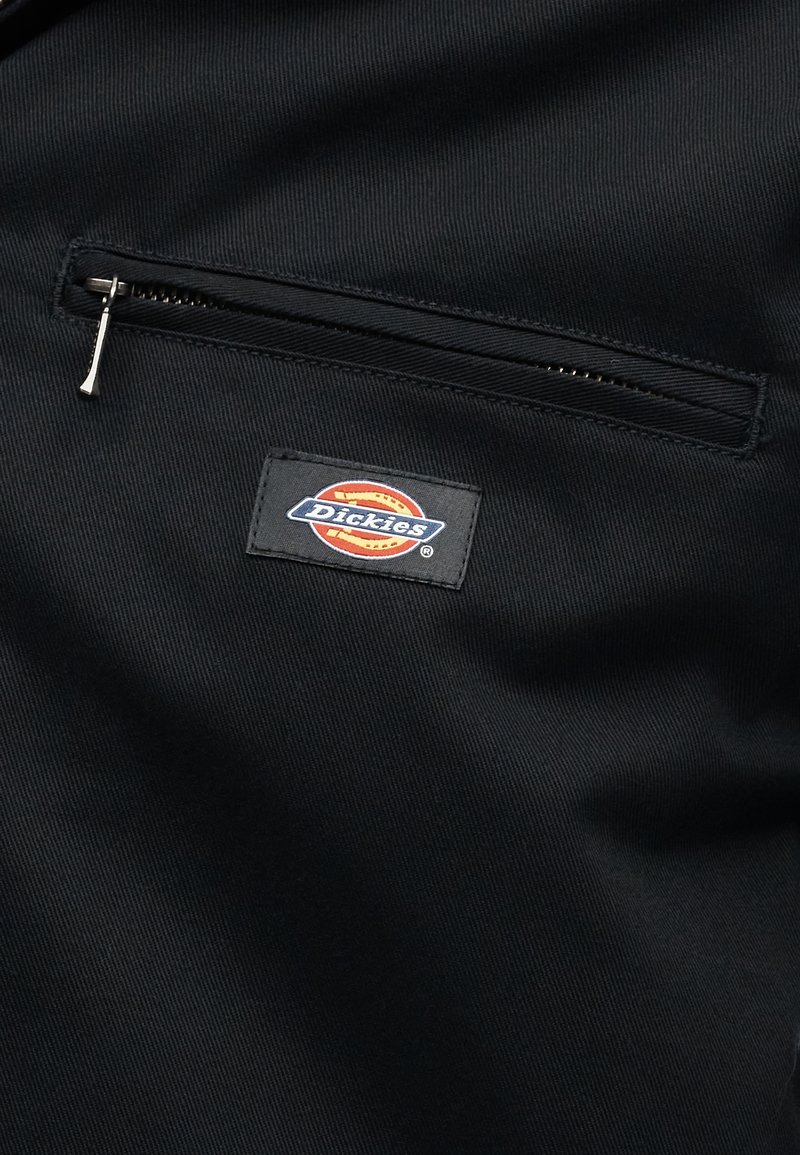 Schwarzer Stoff mit einer horizontalen Reißverschlusstasche oberhalb eines rechteckigen Dickies-Logopatches in Rot-, Orange- und Blautönen.