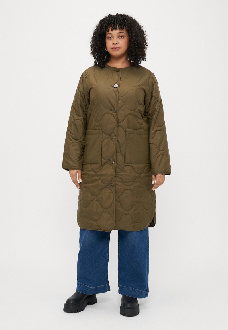 Ελαιόλαδο πράσινο, επενδυμένο μακρύ παλτό με στρογγυλή λαιμόκοψη, κούμπωμα με κουμπιά και δύο μπροστινές τσέπες. Φορεμένο πάνω από γαλάζια παντελόνια με φαρδιά πόδια και μαύρες μπότες.