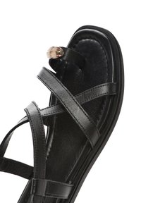 Sandalo in pelle nera con cinturini incrociati e un accento metallico rotondo. Suola piatta con una superficie liscia e un design minimalista.