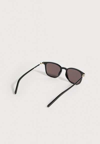 Μαύρα ορθογώνια γυαλιά ηλίου με σκούρους φακούς και λεπτά μπράτσα που ακουμπούν σε ανοιχτή επιφάνεια.