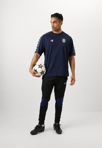 T-shirt à manches courtes bleu marine avec logo et rayures latérales à motifs, associé à un pantalon de survêtement noir avec des accents bleus. Tenant un ballon de football.