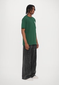 Green cotton t-shirt with a chest pocket, paired with loose-fit black denim pants. Model stands sideways, wearing light-colored sneakers.