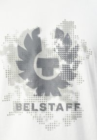 Pilkas Bellstaff prekės ženklas su stilizuota sparnu ir tinklelio raštais ant balto audinio, tekstas "BELLSTAFF" po piešiniu.