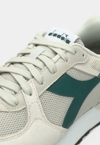 Chaussure de sport beige clair avec une tige en mesh, des accents en daim, un logo bleu sarcelle foncé et des lacets plats. Panneaux texturés près de la zone des orteils.