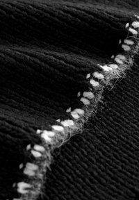 Schwarzer Strickstoff mit einer strukturierten Oberfläche, der am Rand mit einem weißen genähten Akzent versehen ist. Das Material wirkt weich und dicht.