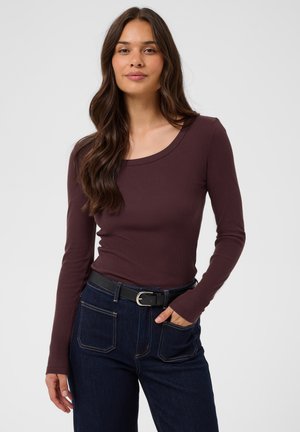 Jeune femme aux longs cheveux ondulés portant un haut marron ajusté à manches longues et un jean bleu foncé avec une ceinture noire, posant avec une main dans la poche.