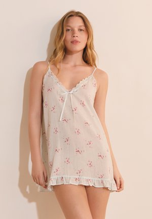 Jonge vrouw met licht haar die een witte mouwloze nachthemd met bloemenprint, kanten afwerking en een strik aan de voorkant draagt, staand tegen een lichte achtergrond.